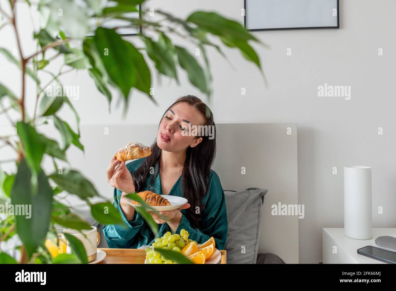 Jeune femme en pyjama de soie a un petit déjeuner sain tout en étant allongé sur le lit dans la chambre. Concept de bien-être à la maison. Santé émotionnelle d'une jeune femme Banque D'Images
