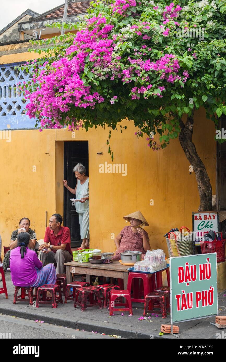 Femme vietnamienne vendant de la nourriture de rue au café de bord de route sous bougainvilliers fleurs, Hoi an, Vietnam Banque D'Images