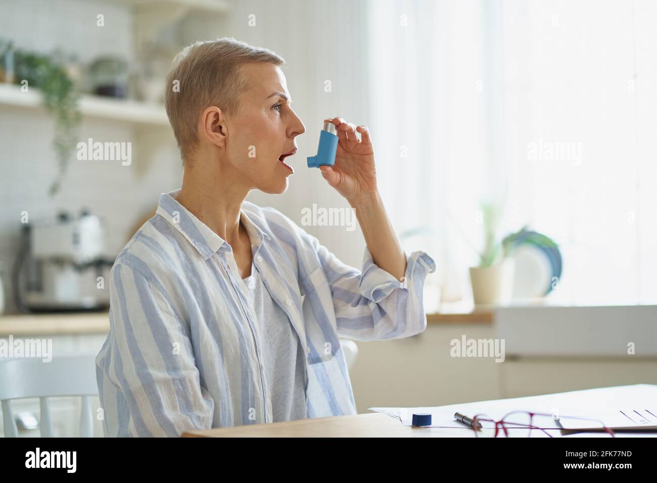 Vue latérale d'une femme d'âge moyen en tenue décontractée en utilisant un inhalateur tout en ayant l'asthme attaque, assis à table dans la cuisine à la maison, l'accent sélectif sur les femmes âgées. Condition médicale et concept de soins de santé Banque D'Images