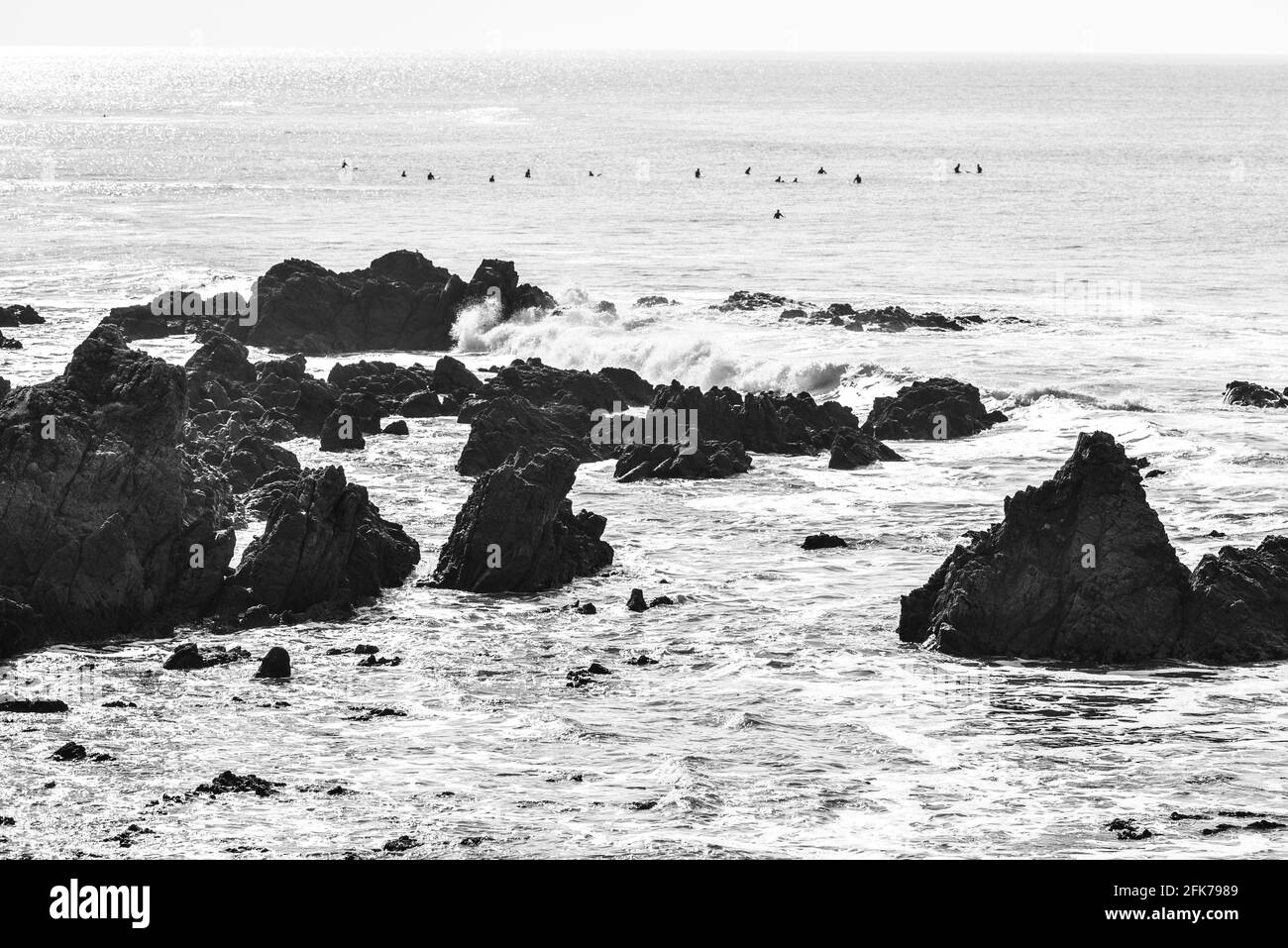 Photo noir et blanc de l'océan et des rochers, avec des surfeurs en arrière-plan attendant une vague. Banque D'Images