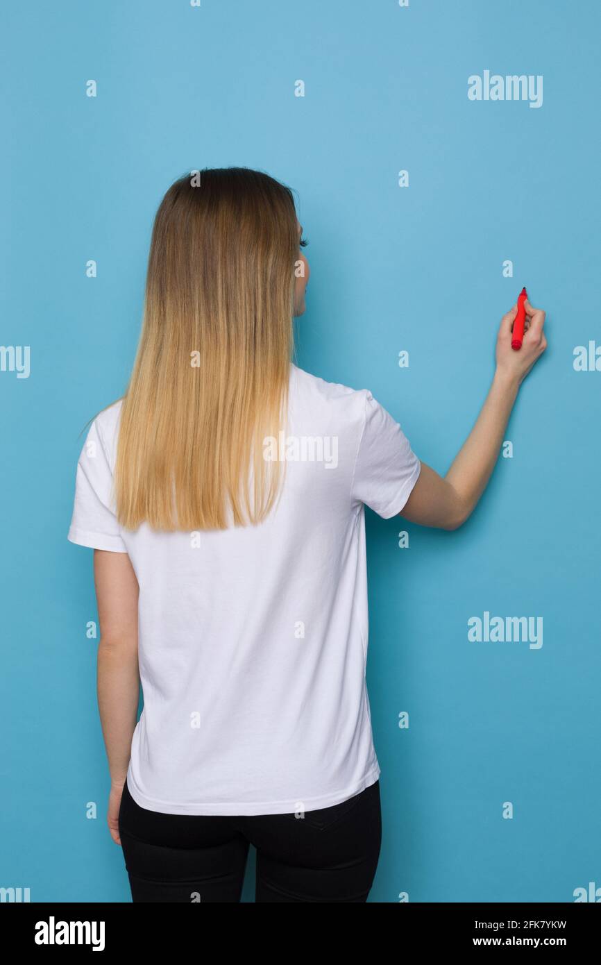 Vue arrière de la jeune femme blonde en chemise blanche, écriture sur un mur bleu avec un stylo rouge. Trois quarts de temps en studio. Banque D'Images