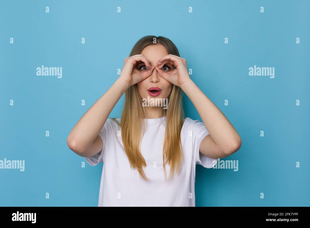 Une jeune femme surprise en chemise blanche fait des jumelles mains et regarde l'appareil photo. Taille haute studio tourné sur fond bleu. Banque D'Images