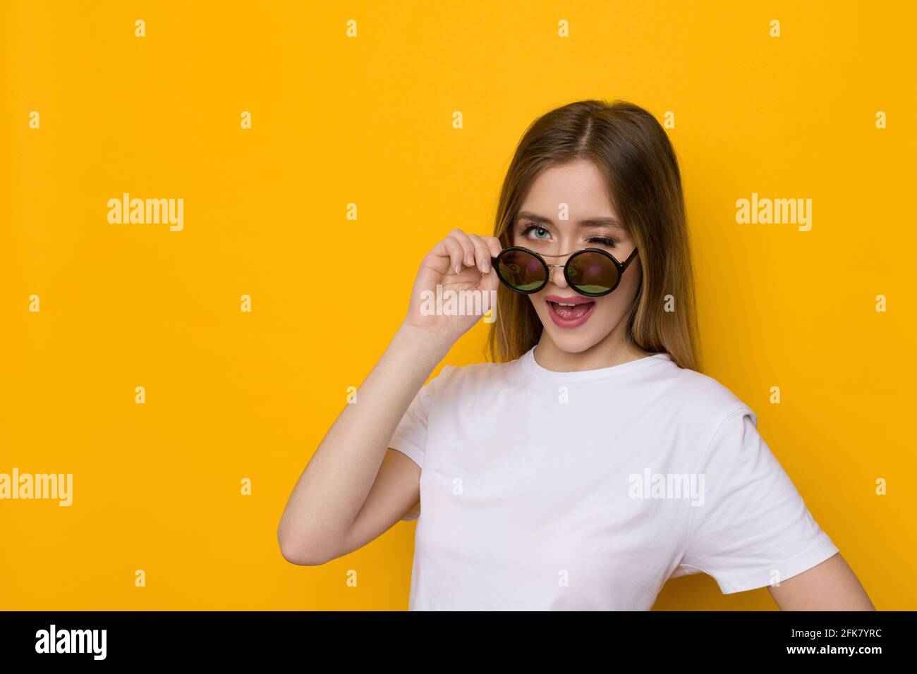 Curieuse jeune femme blonde en chemise blanche est regarder loin au-dessus de lunettes de soleil et de wencrage. Taille haute studio tourné sur fond jaune. Banque D'Images