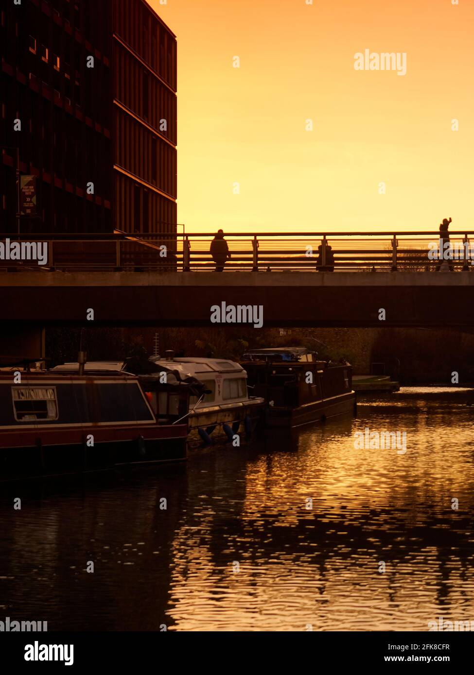 Les silhouetés traversent un pont au-dessus d'un canal urbain, avec des bateaux étroits en dessous et de nouveaux bâtiments derrière. L'eau du canal reflète un ciel doré Banque D'Images