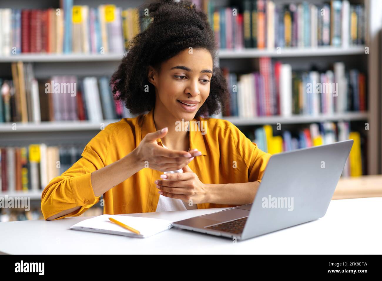 Communication en ligne. Confiante et attrayante fille afro-américaine élégante, étudiante dans une bibliothèque universitaire utilise un ordinateur portable, parlant sur un ami de vidéoconférence ou un camarade de classe, se faisant un geste de la main Banque D'Images