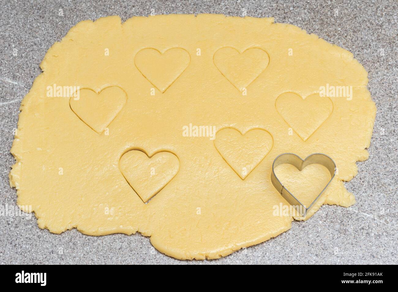 Biscuits coupés au coeur et forme de coeur pour couper sur la pâte jaune crue sur un fond de table gris. Étalez la pâte et découpez les biscuits en forme de coeur pour cuire Banque D'Images