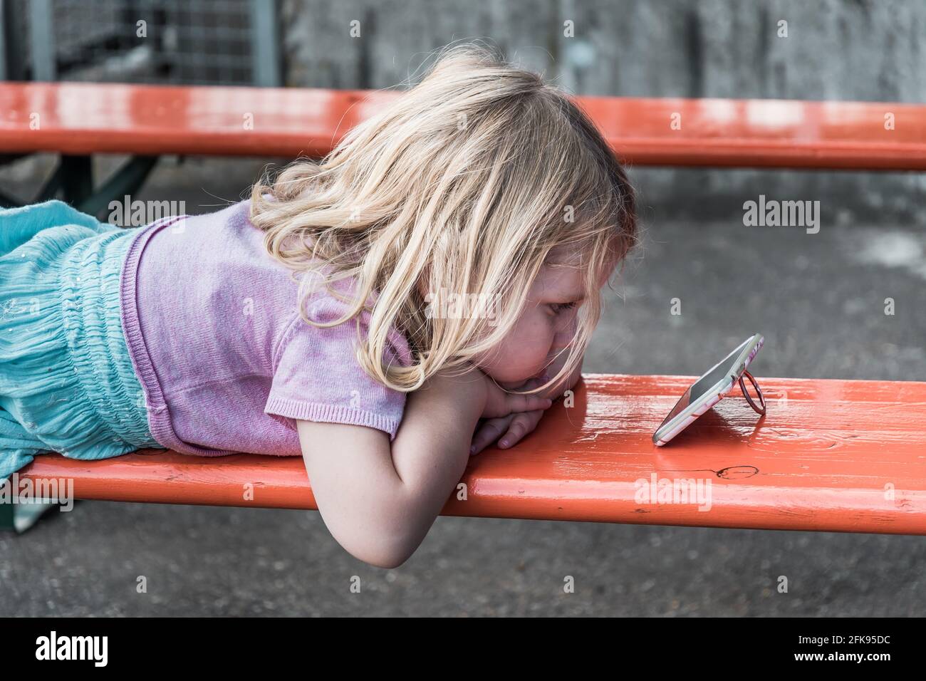 Petite fille blonde est sur un banc regardant une vidéo sur un smartphone Banque D'Images