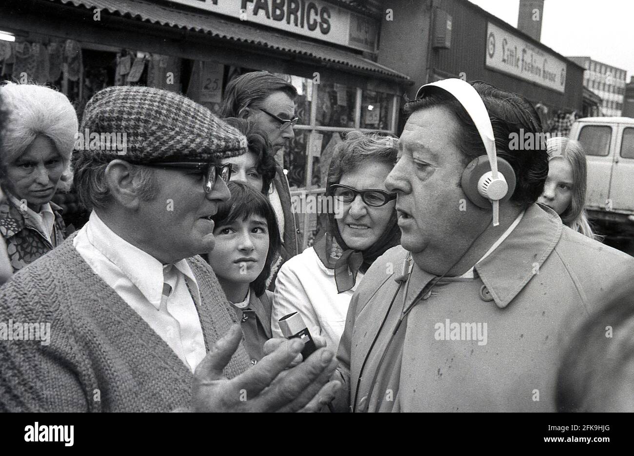 Années 1970, homme populaire du peuple, présentateur de radio et  journaliste, Monty Modlyn parlant à un homme local portant une casquette  plate entouré par des gens dans une zone de marché du