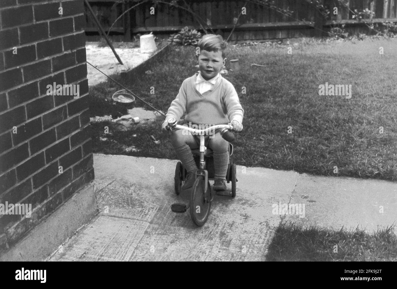 1966, historique, à l'extérieur sur un chemin dans un jardin à l'arrière, à l'angle d'une maison, un petit garçon assis sur son tricycle. Un bac à sable peut également être vu, Angleterre, Royaume-Uni. Banque D'Images