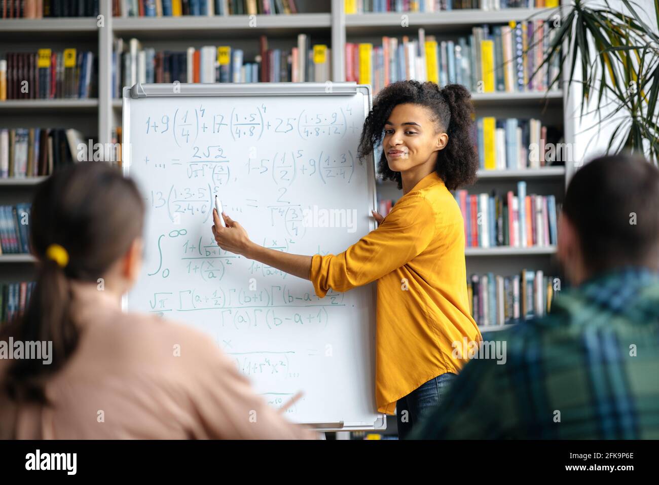 Des élèves attentifs et intelligents écoutent une jeune femme enseignante de race mixte debout sur un tableau blanc, portant des vêtements élégants, elle mène des leçons, fait des brainstorming, montre des informations et sourit Banque D'Images