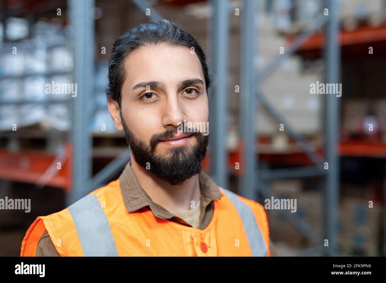 Portrait souriant de jeune ouvrier mixte de race orange gilet debout contre des étagères métalliques au magasin Banque D'Images