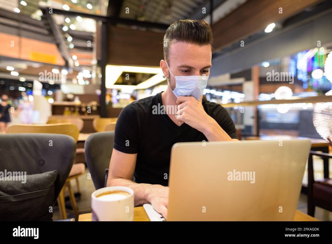 Homme assis au café portant un masque et utilisant ordinateur portable tout en pensant Banque D'Images