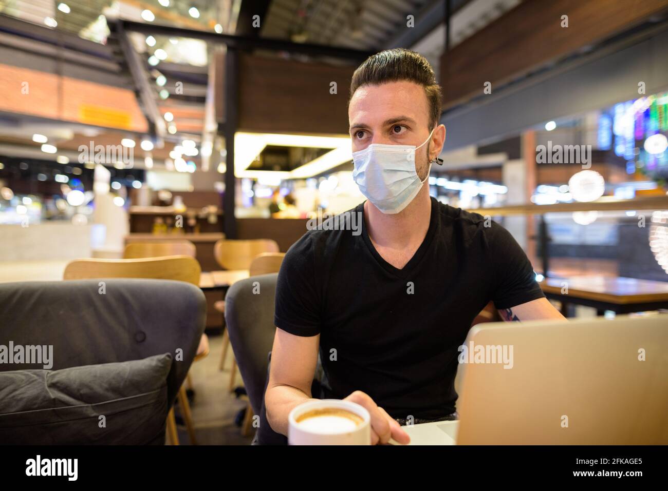 Homme sérieux assis au café-restaurant portant un masque facial et utiliser un ordinateur portable tout en pensant Banque D'Images