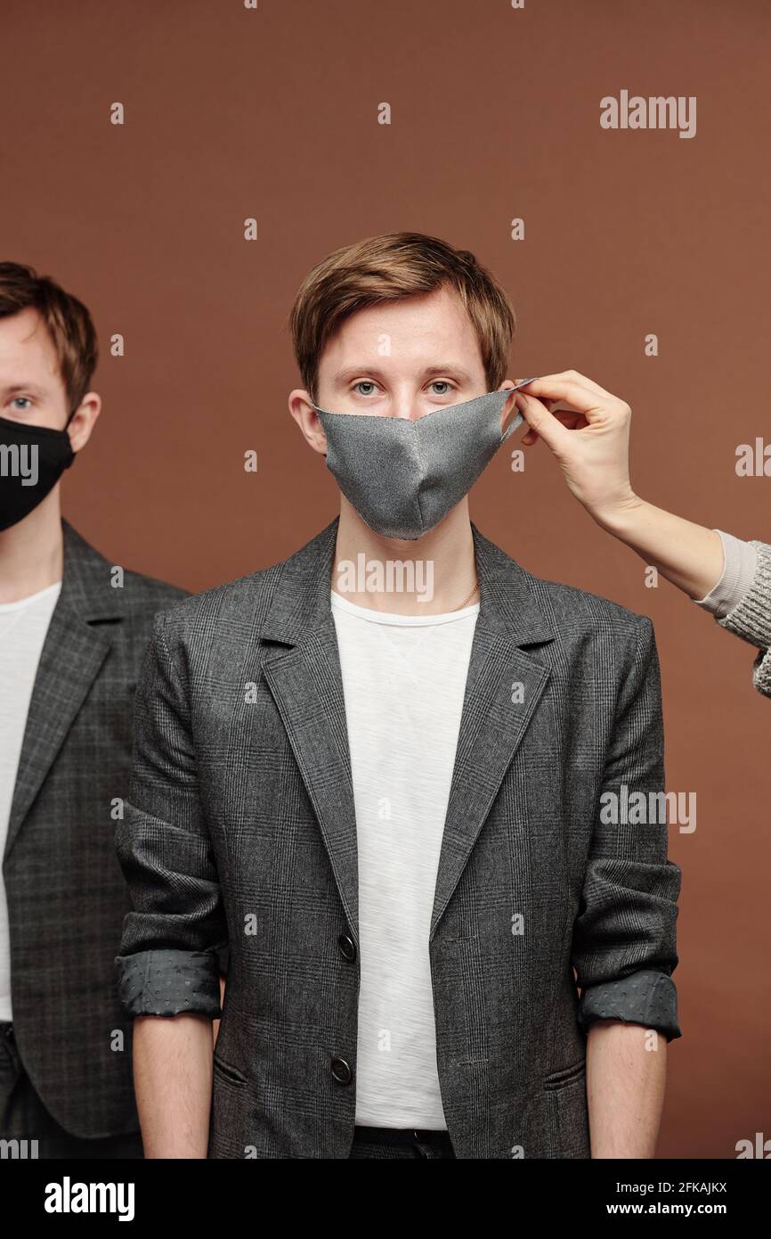 Main mettant le masque sur l'oreille de jeune gars en gris costume debout avec frère jumeau sur fond marron Banque D'Images