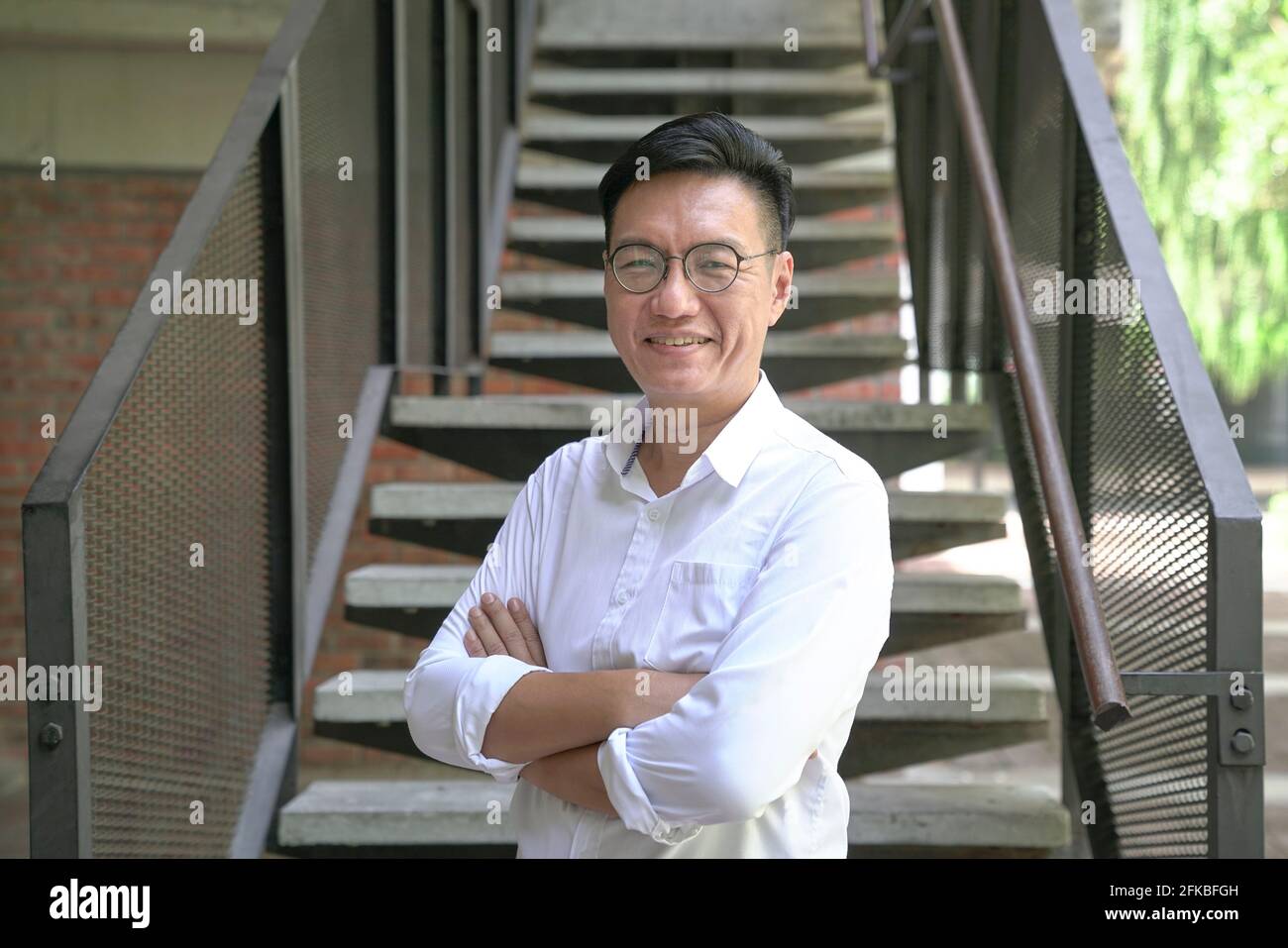 Portrait d'un homme d'affaires asiatique souriant aux bras croisés, portant une chemise blanche. Arrière-plan de l'escalier. Banque D'Images