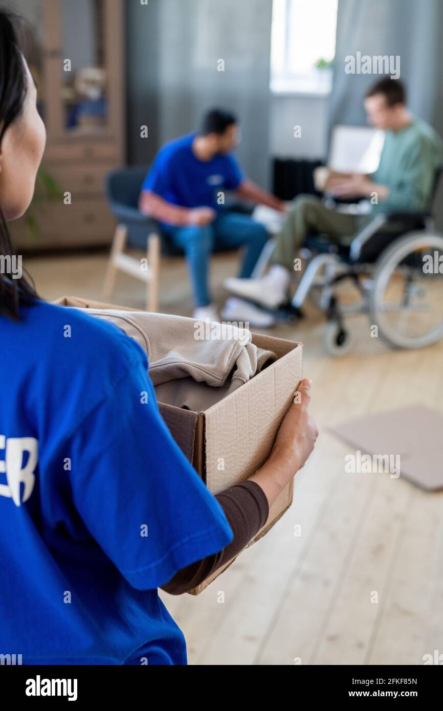 Une femme volontaire porte une boîte avec des vêtements contre l'homme dans un fauteuil roulant et sa collègue Banque D'Images