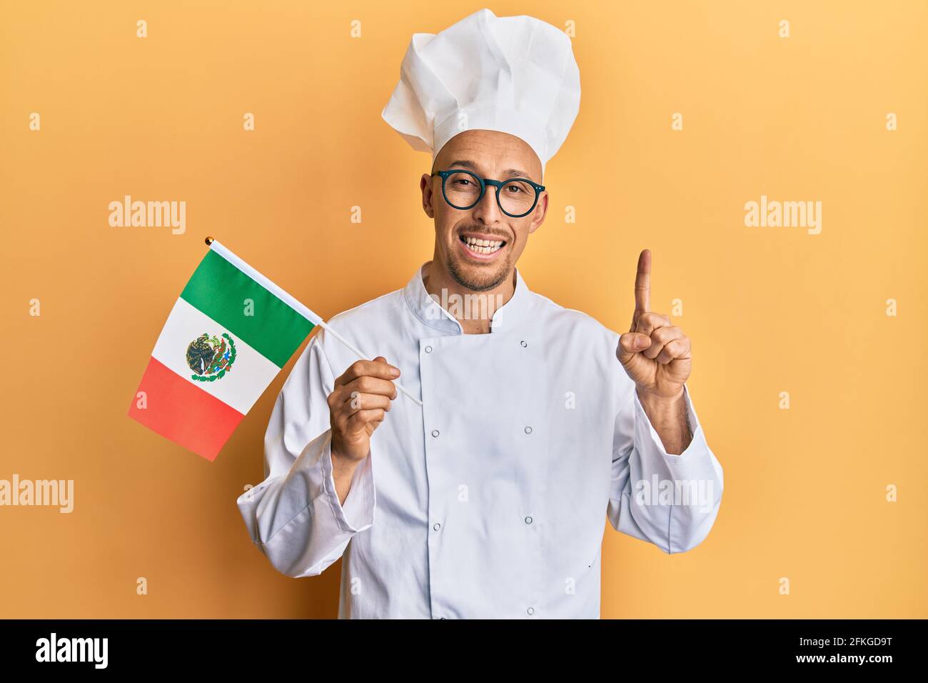Homme chauve avec barbe portant un tablier de cuisinier professionnel tenant le drapeau du mexique souriant avec une idée ou une question pointant du doigt avec le visage heureux, numéro un Banque D'Images