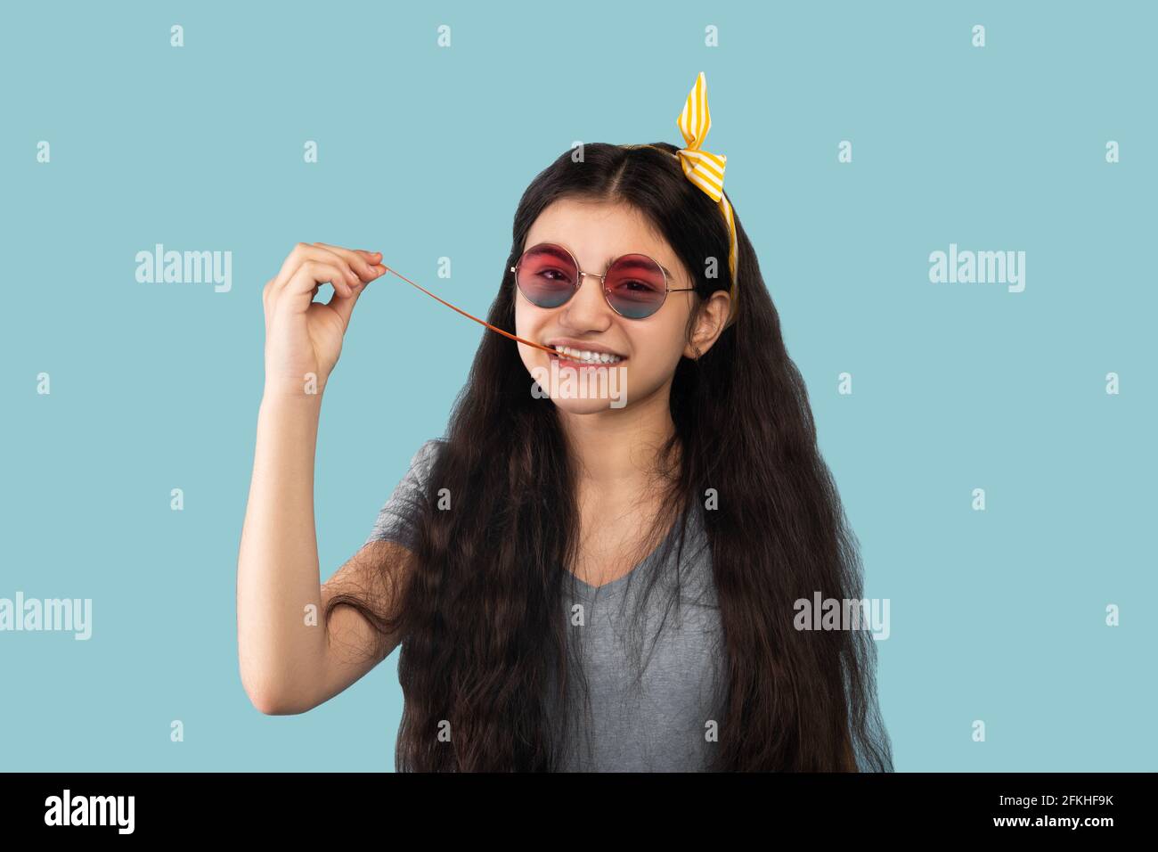 Portrait d'une jeune fille indienne ludique en tenue décontractée et lunettes de soleil prenant de la gomme à bulles hors de sa bouche, fond bleu Banque D'Images