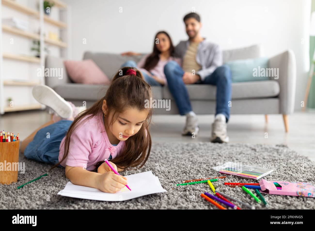 Bonheur de famille. Jolie fille dessin avec des crayons colorés, couché sur le tapis de sol tandis que les parents se détendent sur le canapé Banque D'Images