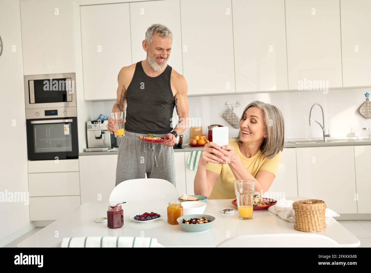 Un couple de familles âgées heureux prenant le petit déjeuner au téléphone dans la cuisine. Banque D'Images