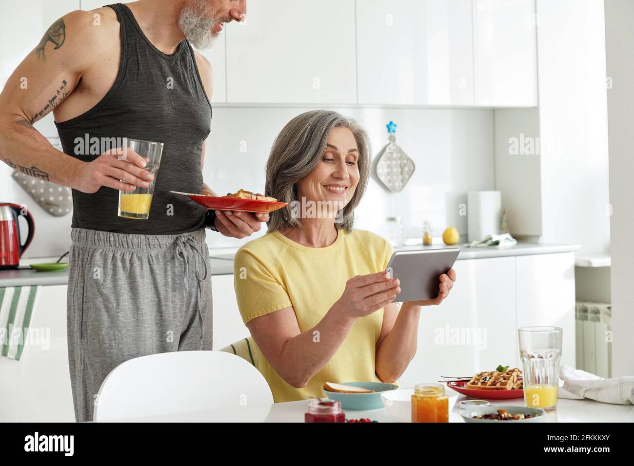 Un couple de familles âgées heureux prenant le petit déjeuner avec une tablette dans la cuisine. Banque D'Images