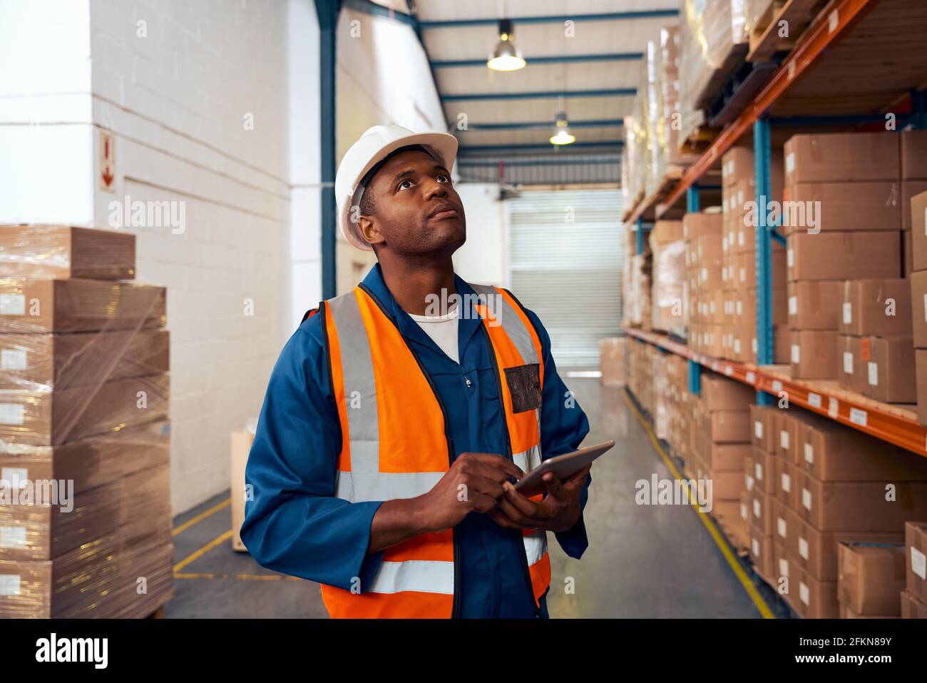Jeune ouvrier d'usine de sexe masculin qui conserve des registres de stock dans un grand entrepôt avec pile de boîtes en carton Banque D'Images