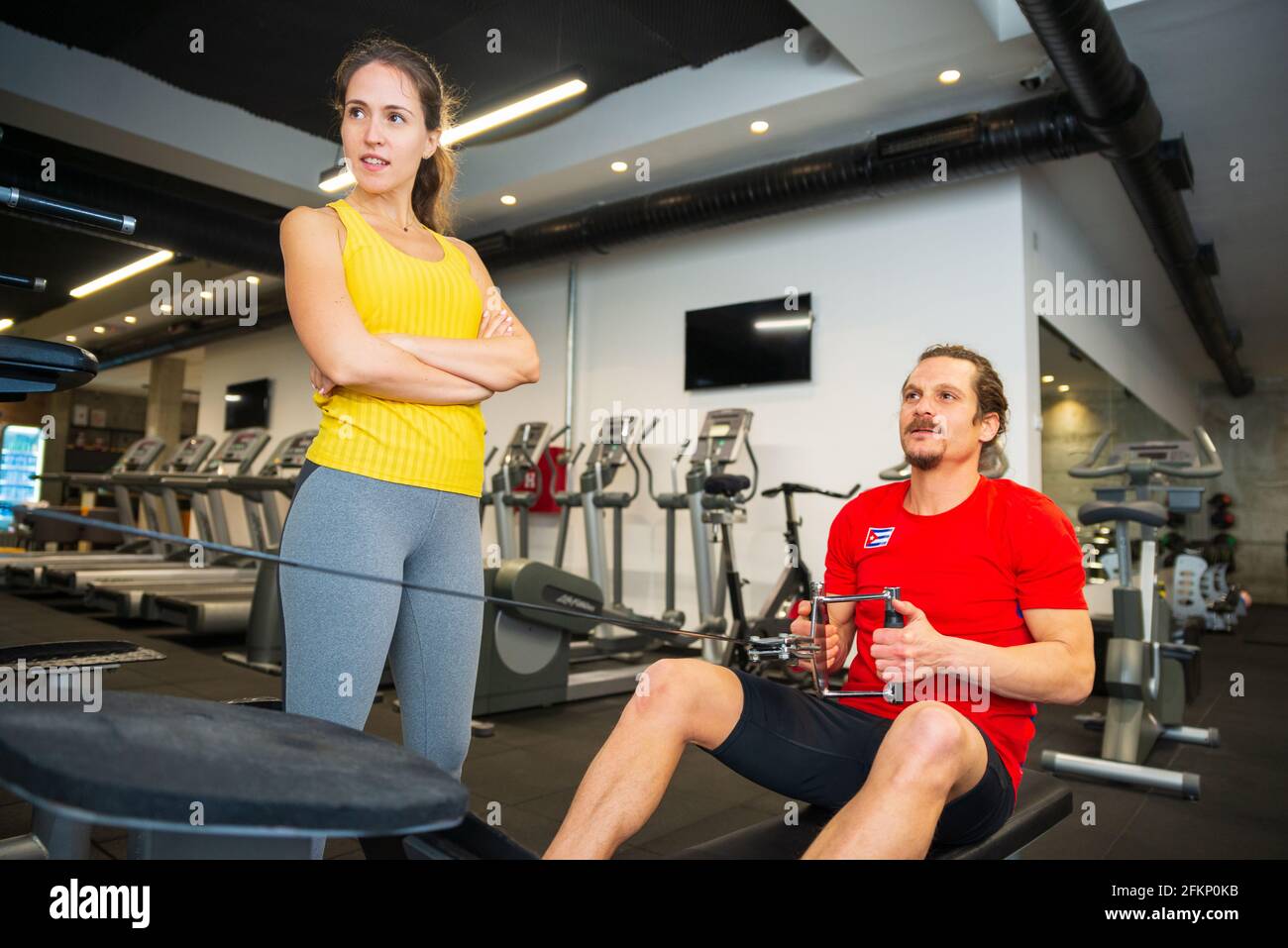 Jeune femme sportive de race blanche debout à côté du jeune homme s'exerçant sur un rameur dans la salle de sport moderne Banque D'Images