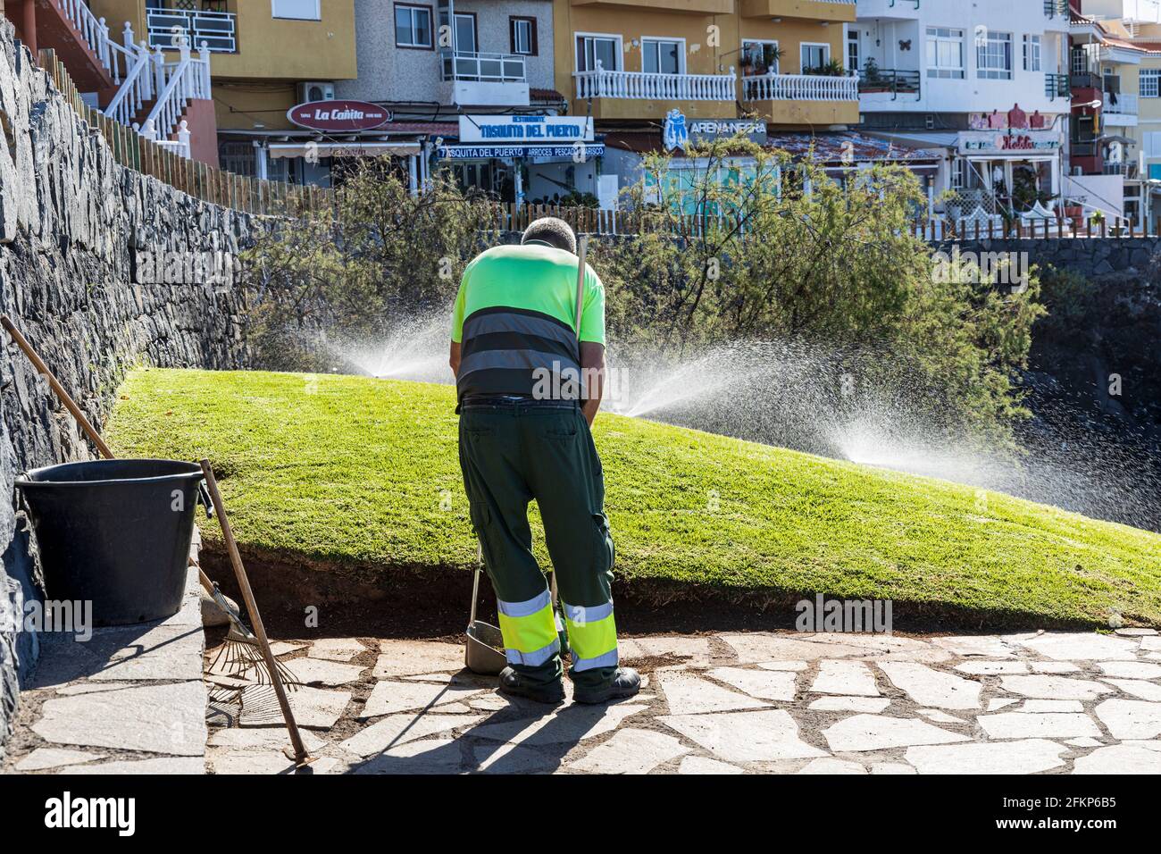 Un travailleur du conseil qui balaie les arroseurs vaporise de l'eau pour irriguer les jardins de Los Abrigos, Tenerife, Iles Canaries, Espagne Banque D'Images