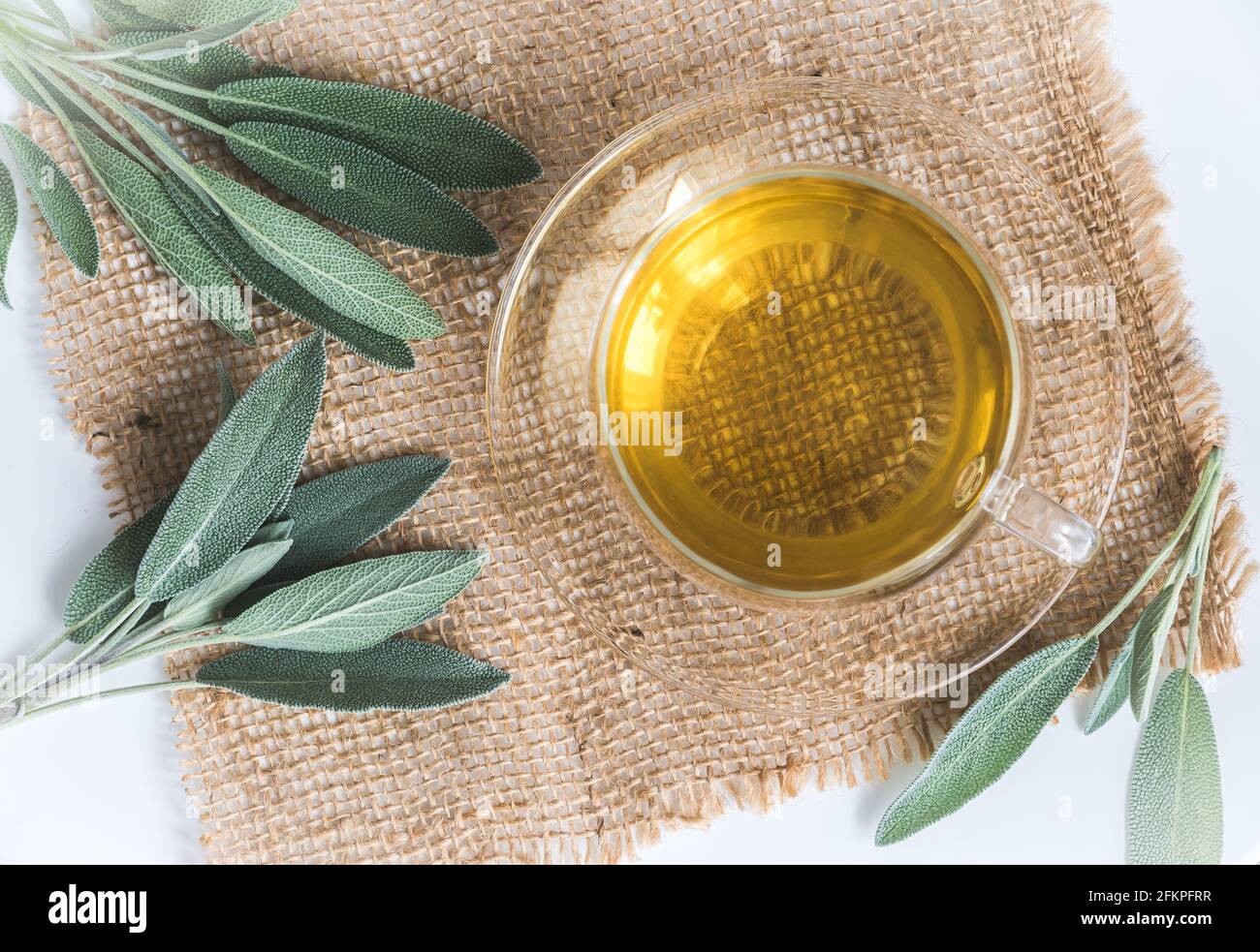 Coupe en verre avec thé et feuilles de sauge sur jute et bois blanc, vue de dessus Banque D'Images