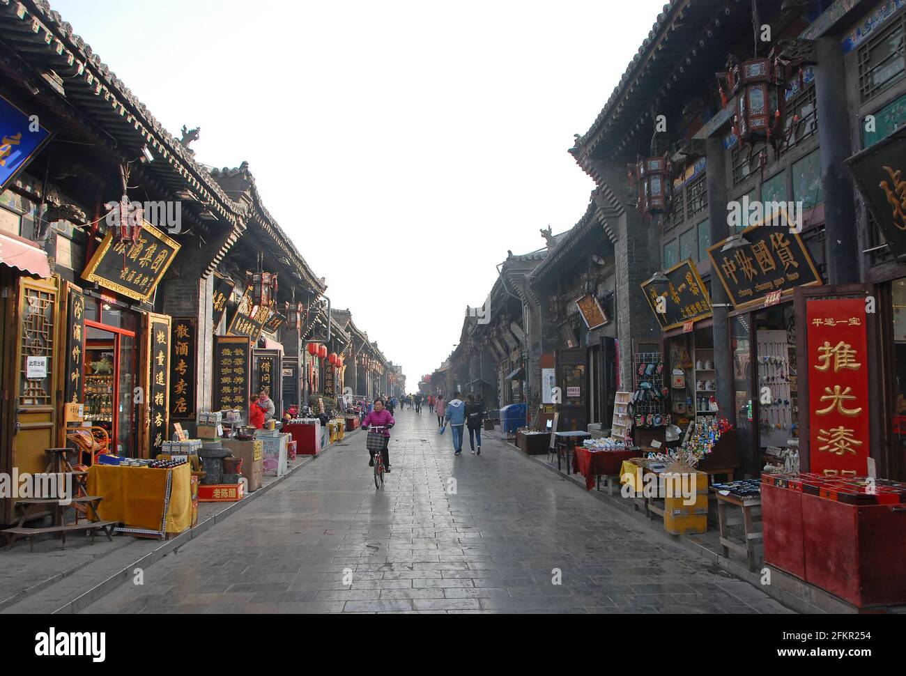 Une rue principale à Pingyao, province du Shanxi, Chine bordée de petits magasins. Une femme fait du vélo et les gens marchent. Banque D'Images