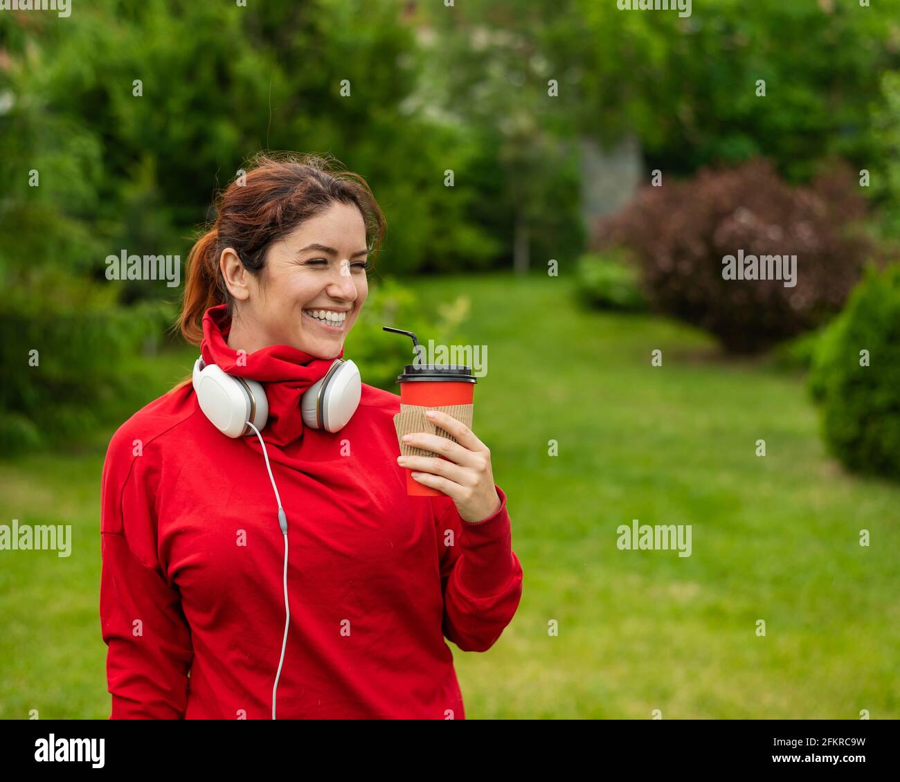 Femme aux cheveux rouges avec casque et boissons café avec une paille dans le parc. Une jeune fille aime se promener par une belle journée d'été et tient un cappuccino à emporter Banque D'Images