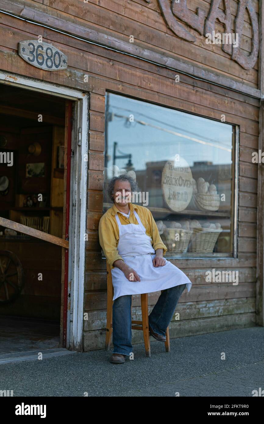 Boulanger souriant assis à l'extérieur près de la porte de sa boulangerie attendant ses clients. Banque D'Images