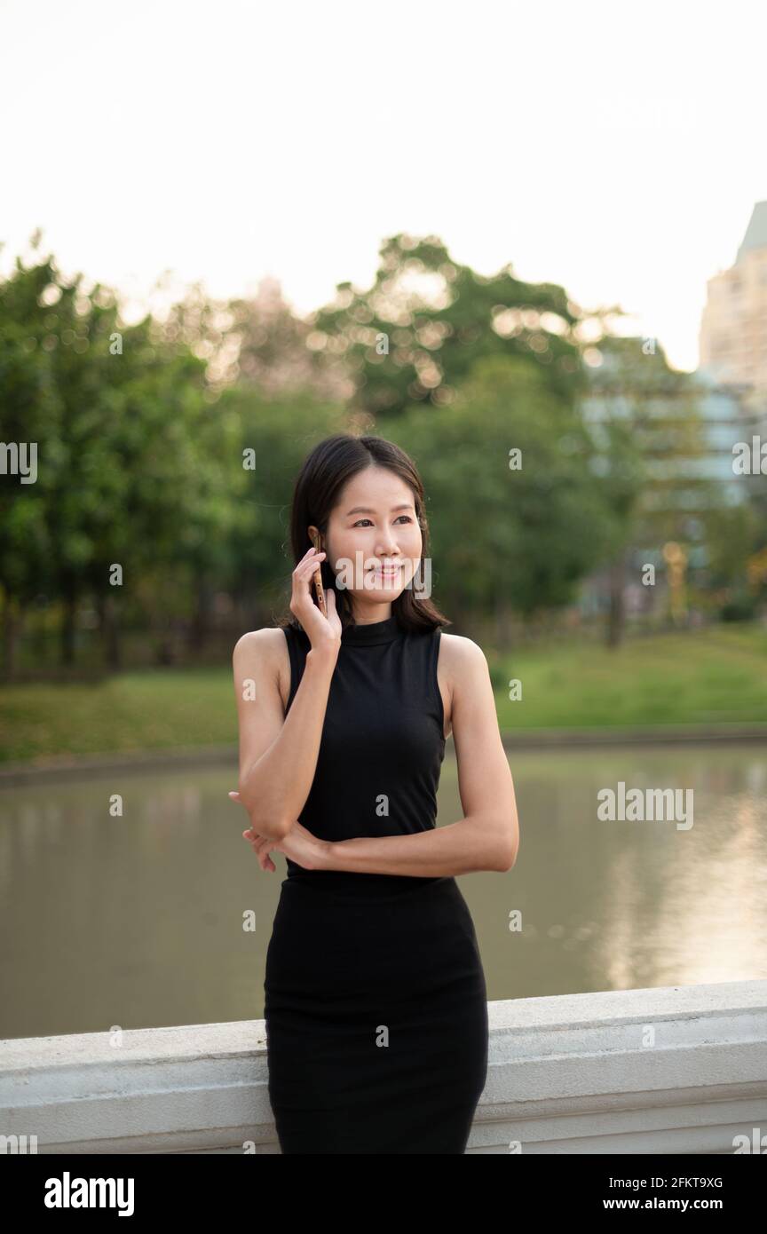 Une jeune femme d'affaires asiatique d'origine ethnique parle sur son téléphone mobile à l'extérieur dans un parc, avec des arbres et des bâtiments en arrière-plan. Banque D'Images