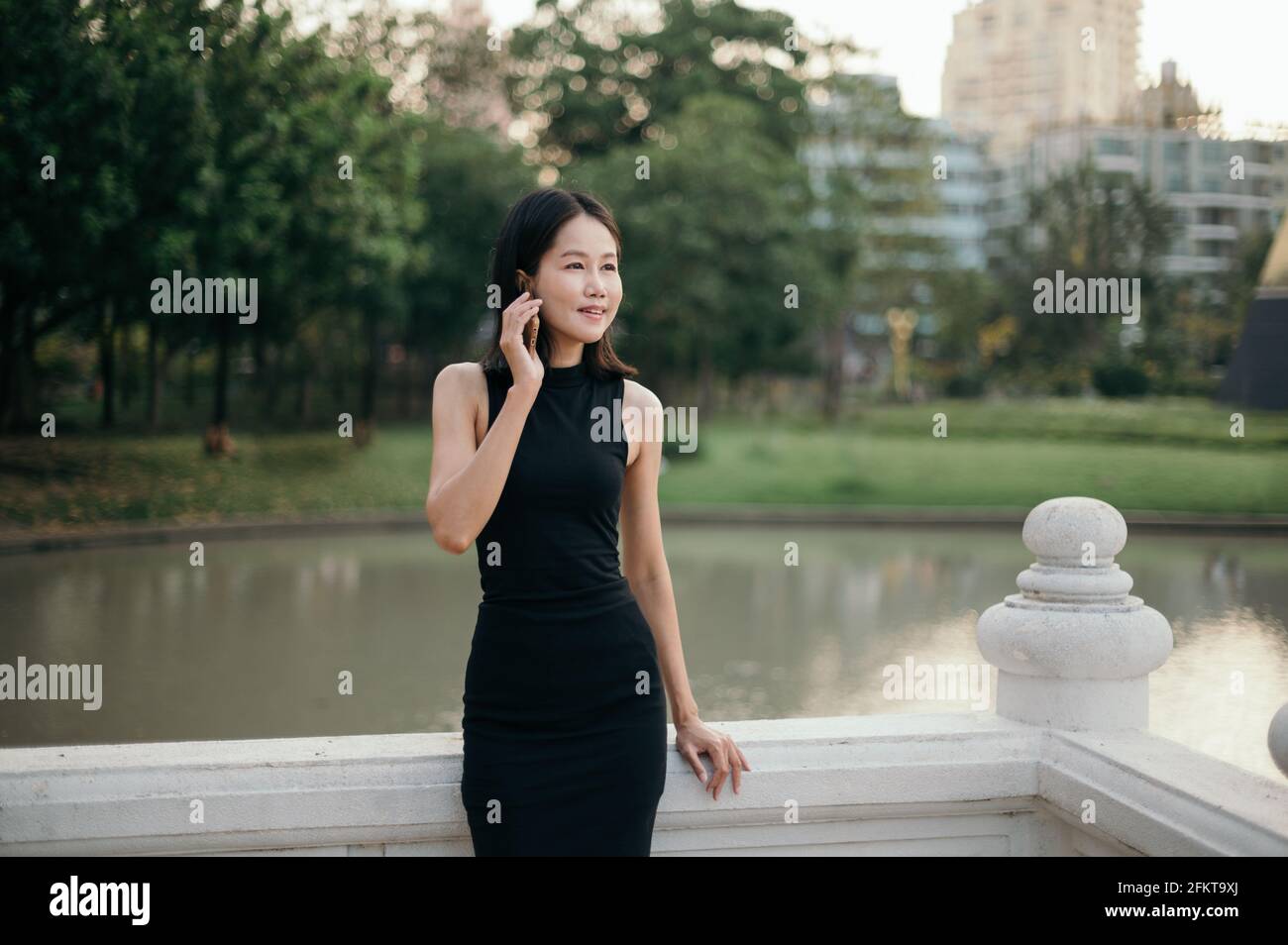Une jeune femme d'affaires asiatique d'origine ethnique parle sur son téléphone mobile à l'extérieur dans un parc, avec des arbres et des bâtiments en arrière-plan. Banque D'Images