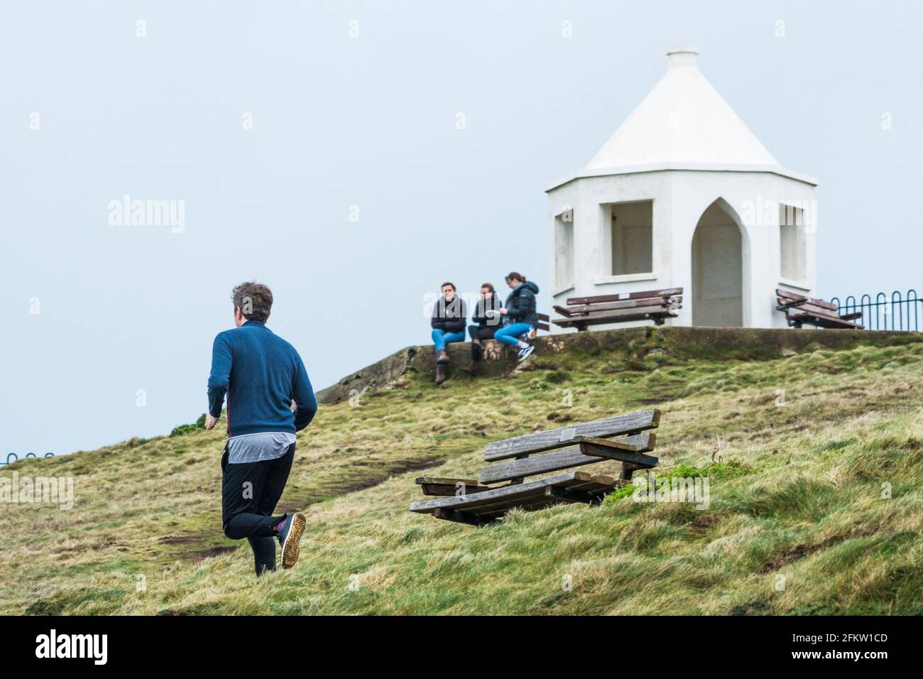 Un homme qui court avec trois femmes assis en arrière-plan sur le dessus de Towan Head à Newquay, en Cornouailles. Banque D'Images