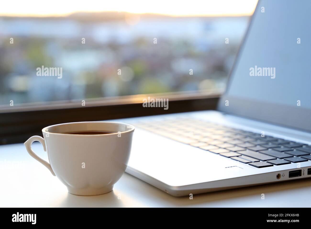 Espace de travail confortable dans le bureau à la maison avec une tasse de café chaude et un ordinateur portable sur la table contre la fenêtre au lever du soleil. Bonjour, concept de travail à distance et de gain Banque D'Images