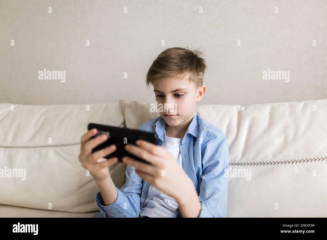 Enfant assis sur le canapé dans ses mains tenant un téléphone, joue avec lui. Banque D'Images