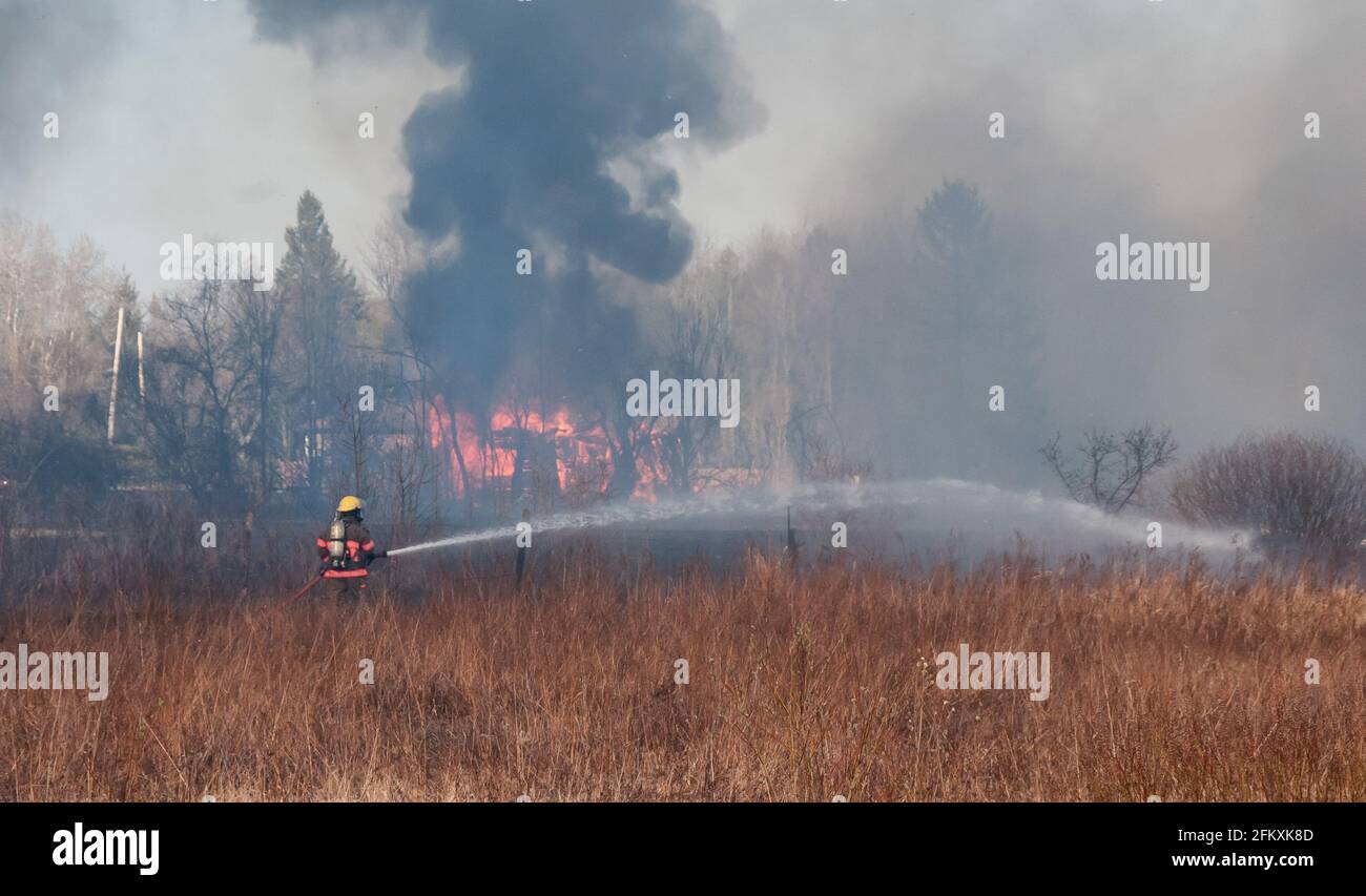 Un pompier vaporisant de l'eau sur de l'herbe brune sèche qui est en feu. Une maison brûle en arrière-plan avec de la fumée noire épaisse. Banque D'Images