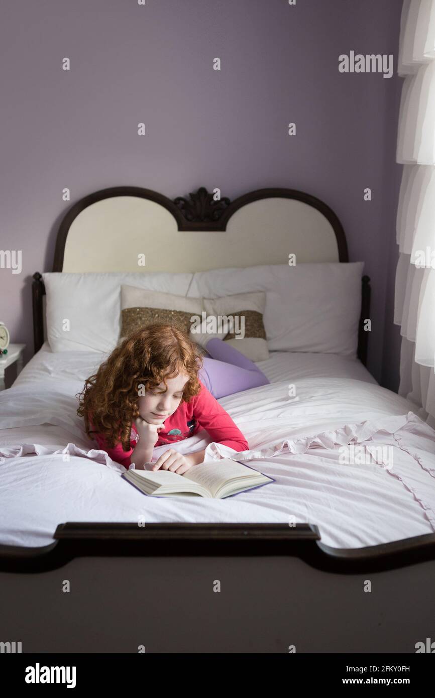 Une jeune fille d'âge scolaire à cheveux rouges lisant sur son lit seul Banque D'Images