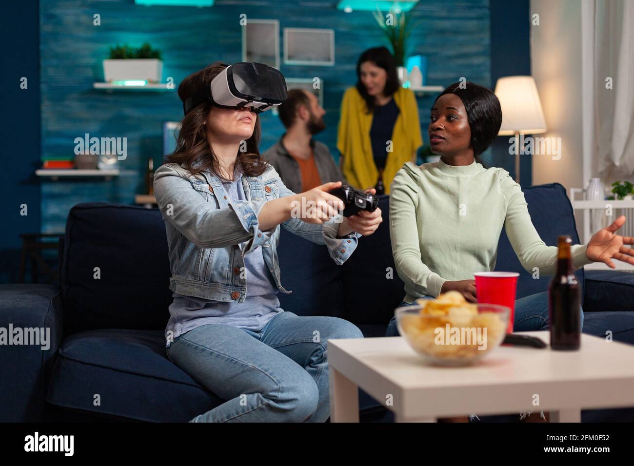 Amis multiethniques soutenant une femme pendant une compétition de jeu vidéo jouant avec des lunettes vr à l'aide d'un joystick. Groupe de course mixte de personnes qui traînaient ensemble en s'amusant tard dans la nuit dans le salon. Banque D'Images