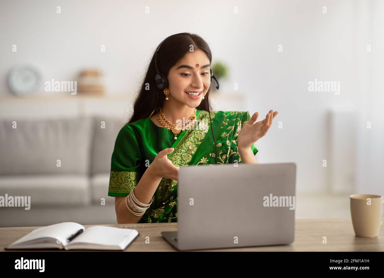 Femme indienne positive en robe traditionnelle sari portant des écouteurs, ayant une réunion en ligne sur ordinateur portable à la maison Banque D'Images