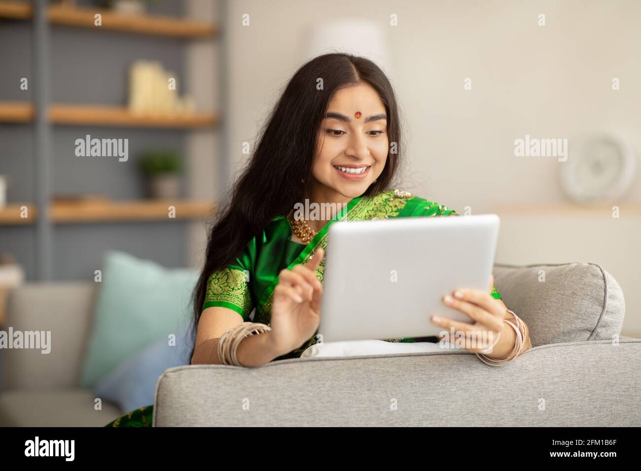 Portrait d'une femme indienne joyeuse en vert sari à l'aide d'une tablette ordinateur sur le canapé à la maison Banque D'Images