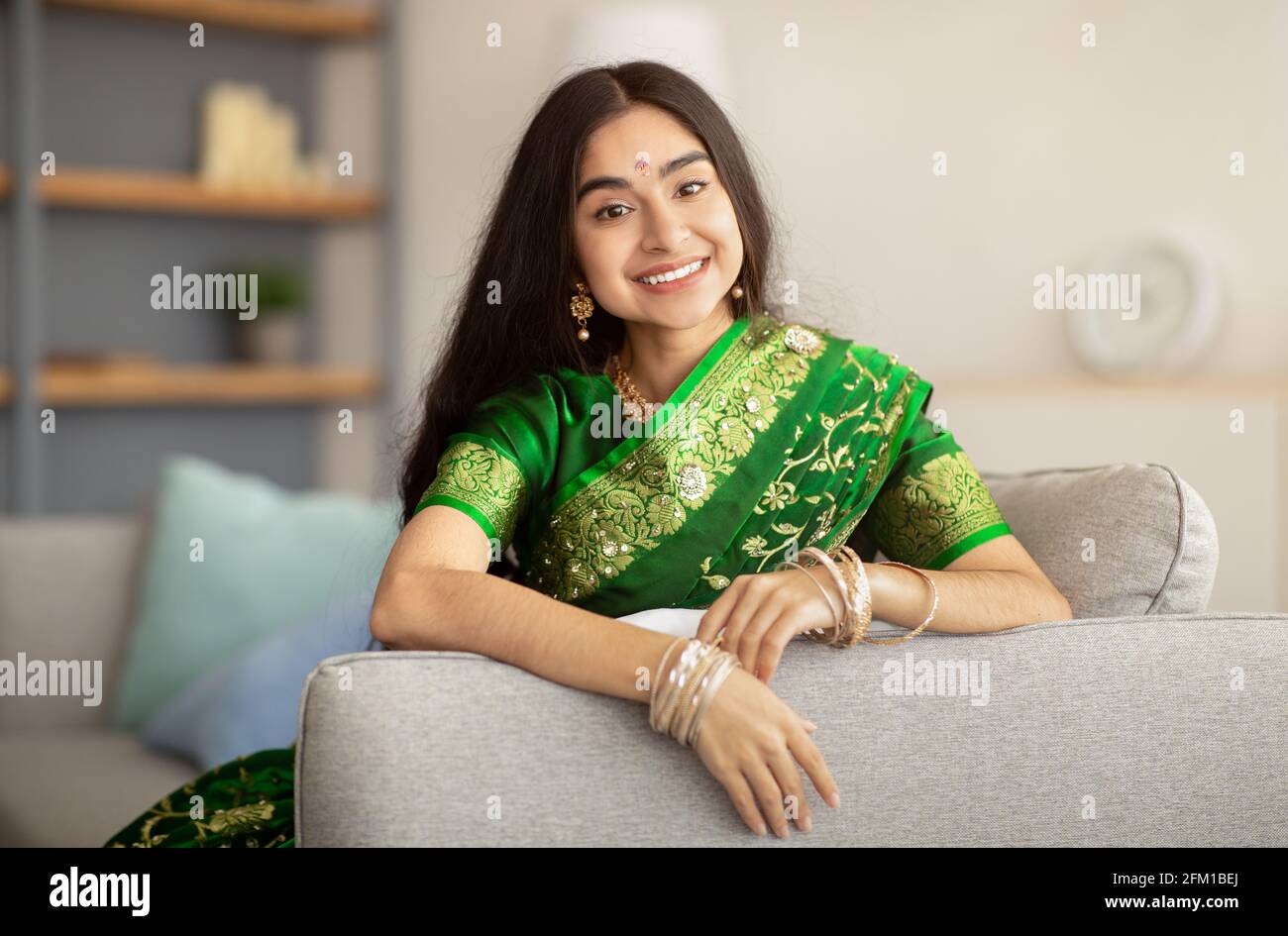 Portrait de dame indienne gaie dans une belle robe verte sari assis sur un canapé et souriant à l'appareil photo, à l'intérieur Banque D'Images