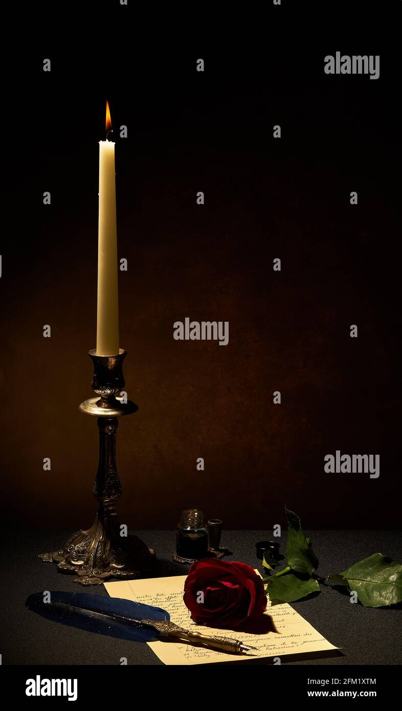 Une bougie en chandelier, rose rouge, lettre et plume stylo sur un fond sombre éclairci par la bougie. Banque D'Images