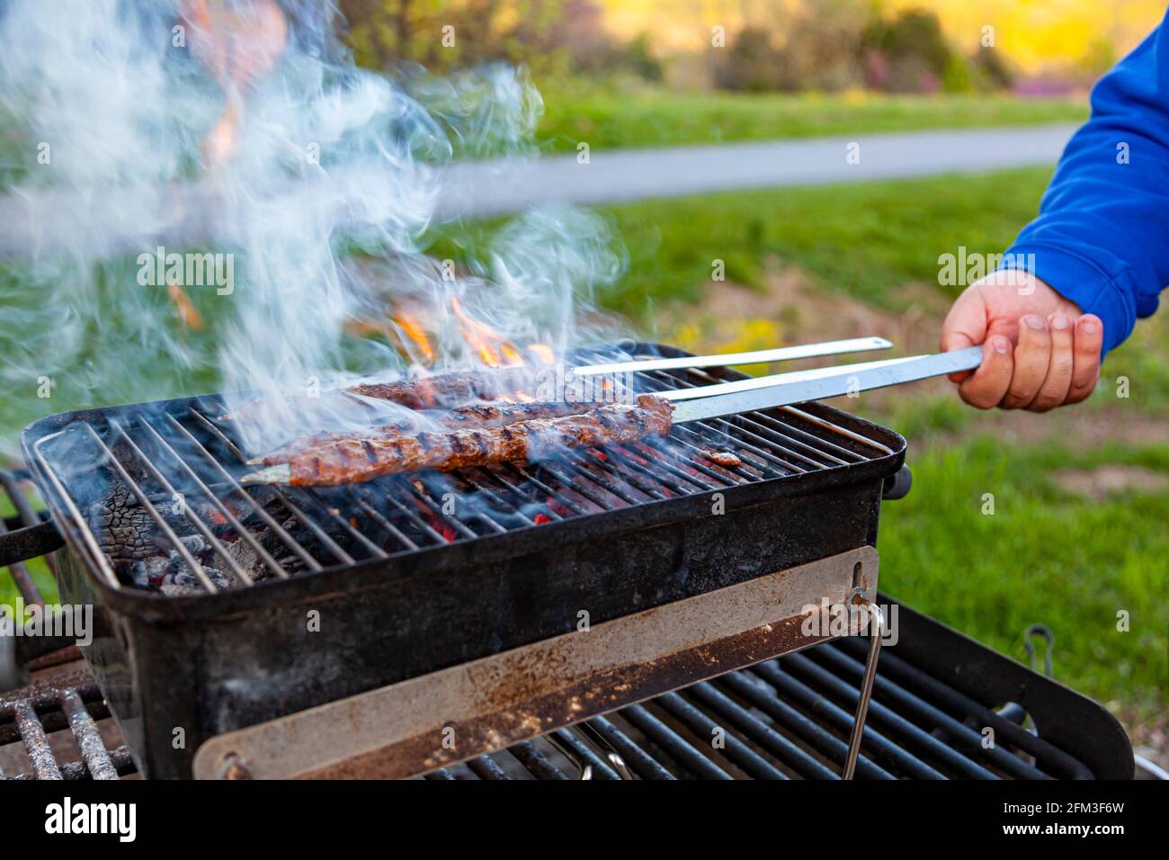 Cuisson de délicieux Adana Kebab turc sur un grill portatif au charbon de bois à un endroit de pique-nique, la fumée et la flamme s'échappe de la viande qui grésilte. Un homme tourne Banque D'Images