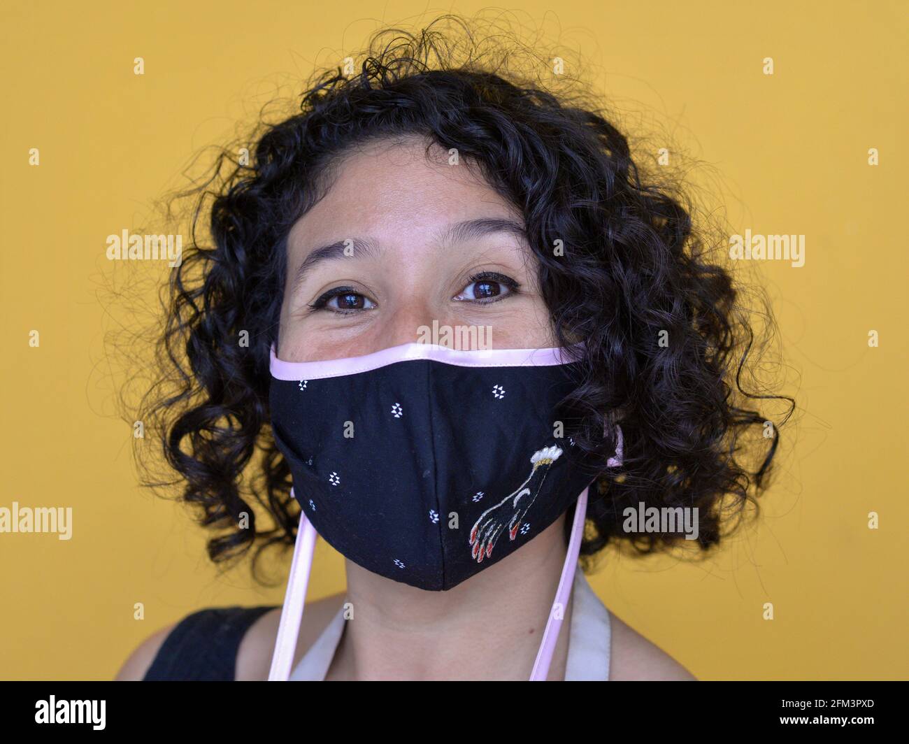 Une jeune femme mexicaine avec des boucles à tire-bouchon porte un masque facial en tissu non médical élégant pendant la pandémie mondiale de coronavirus et regarde la caméra. Banque D'Images