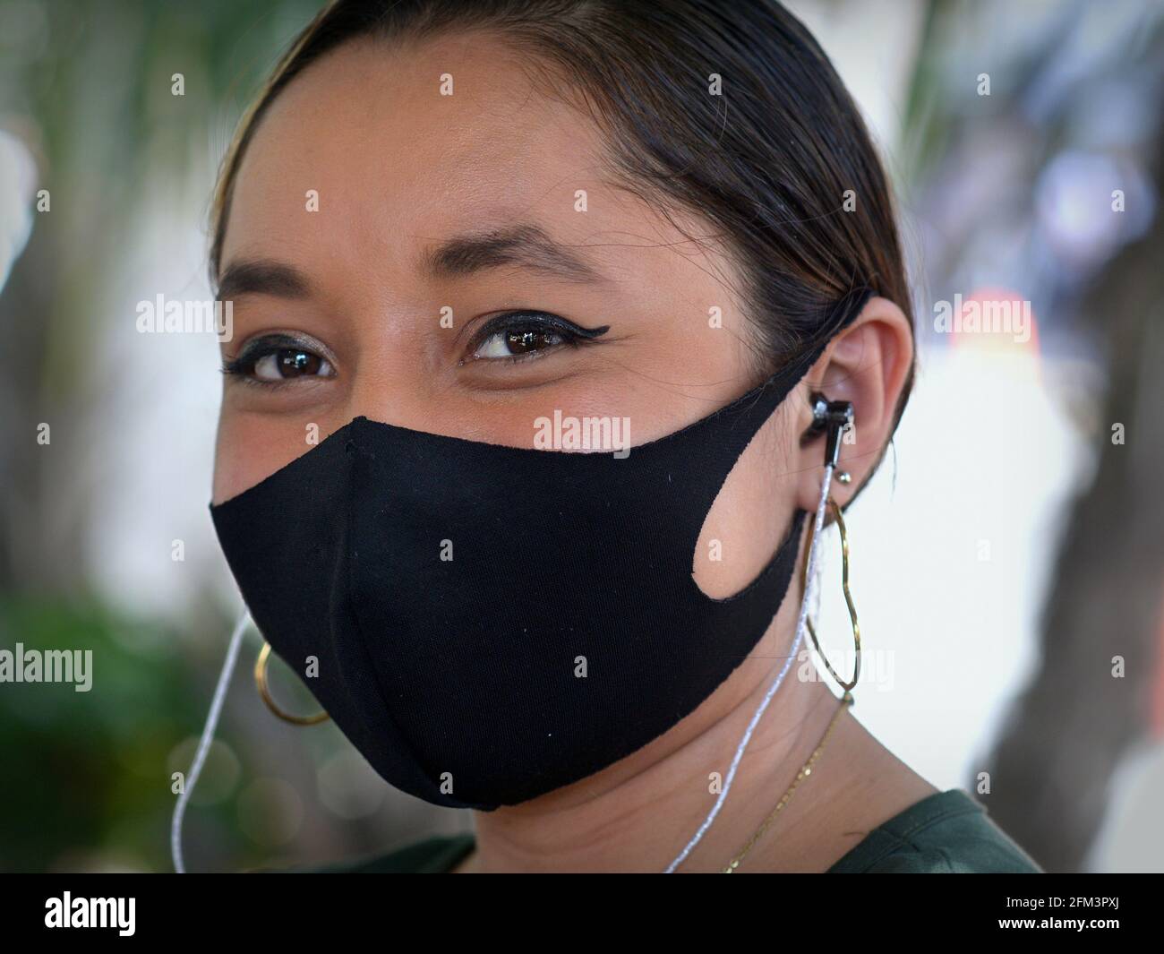 La jeune femme mexicaine optimiste, avec écouteurs, porte un masque noir en tissu non médical pendant la pandémie du coronavirus et sourit avec les yeux. Banque D'Images