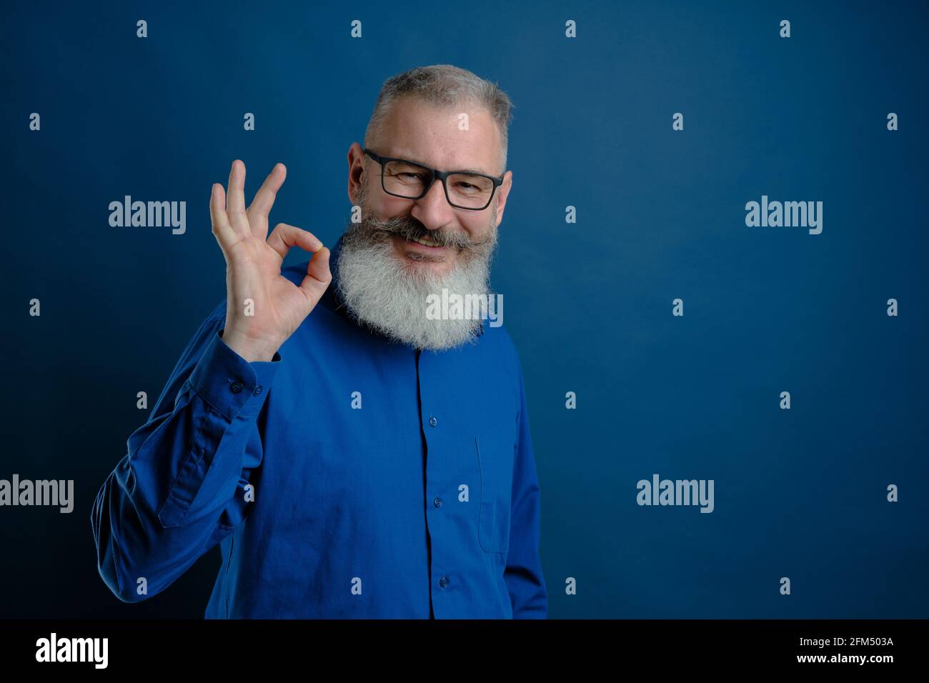 Portrait d'homme caucasien à poil gris mature habillé de chemise bleue montre le signe ok, fond bleu, foyer sélectif Banque D'Images