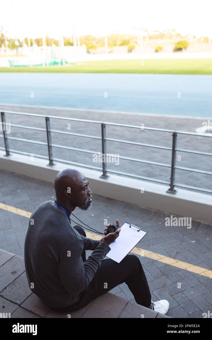 Entraîneur mâle afro-américain avec chronomètre mesurant le temps en position assise sur les sièges dans le stade Banque D'Images