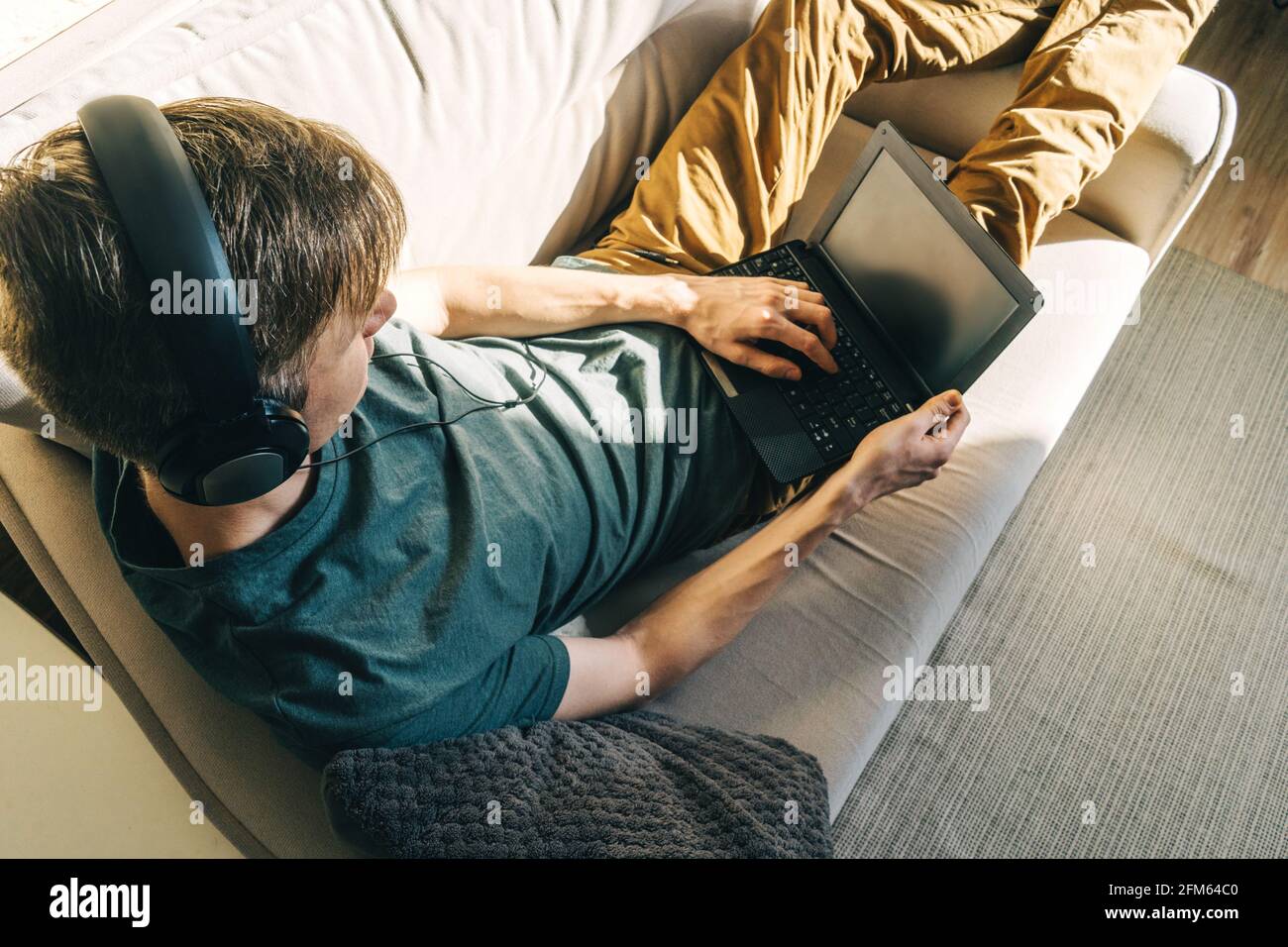 Un jeune homme dans un casque travaille avec un ordinateur portable sur un canapé à la maison. Vue de dessus, jour ensoleillé. Banque D'Images